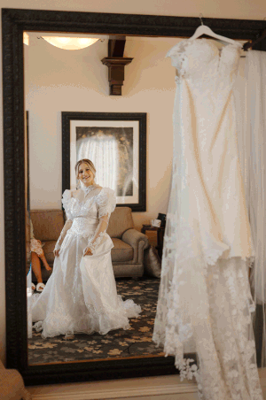 A reflection in a large mirror shows an elegant white lace wedding dress with another gown hanging beside it.