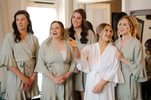 A group of bridesmaids in sage green dresses laugh and smile together while getting ready for a wedding.