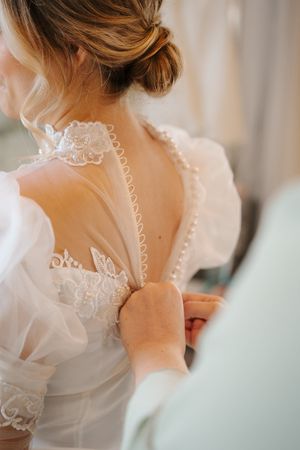 Close up view of delicate white lace and tulle wedding dress being buttoned up, showing intricate floral details.