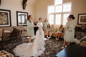 A bride in a flowing white wedding dress gets ready with her bridal party in a sunlit room.