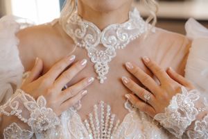 Close-up details of delicate white lace and embroidery on wedding dress with hands showing elegant manicure.
