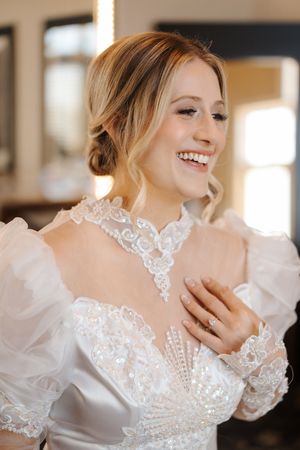 A bride wearing an elegant white lace wedding dress with sheer puff sleeves smiles joyfully on her special day.