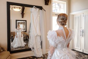 A white wedding dress hangs on display next to a mirror while a bride stands in her gown in a sunlit room.