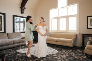 A wedding party gets ready in a sunlit room with beige walls and large windows.
