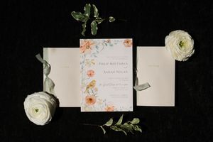 Wedding invitation suite with floral design laid flat on black background with white ranunculus flowers and greenery.