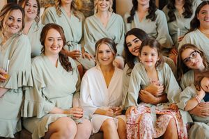 A group of people wearing light colored robes and dresses gather together in a bridal preparation setting.