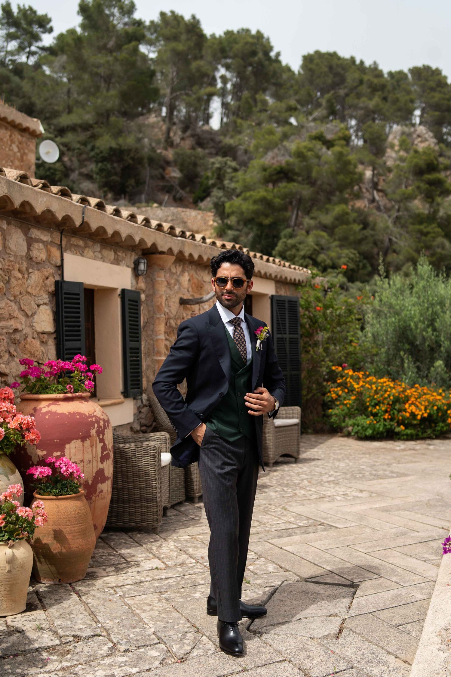 Groom in dark suit and sunglasses poses on stone terrace with colorful flowers and mountain backdrop.