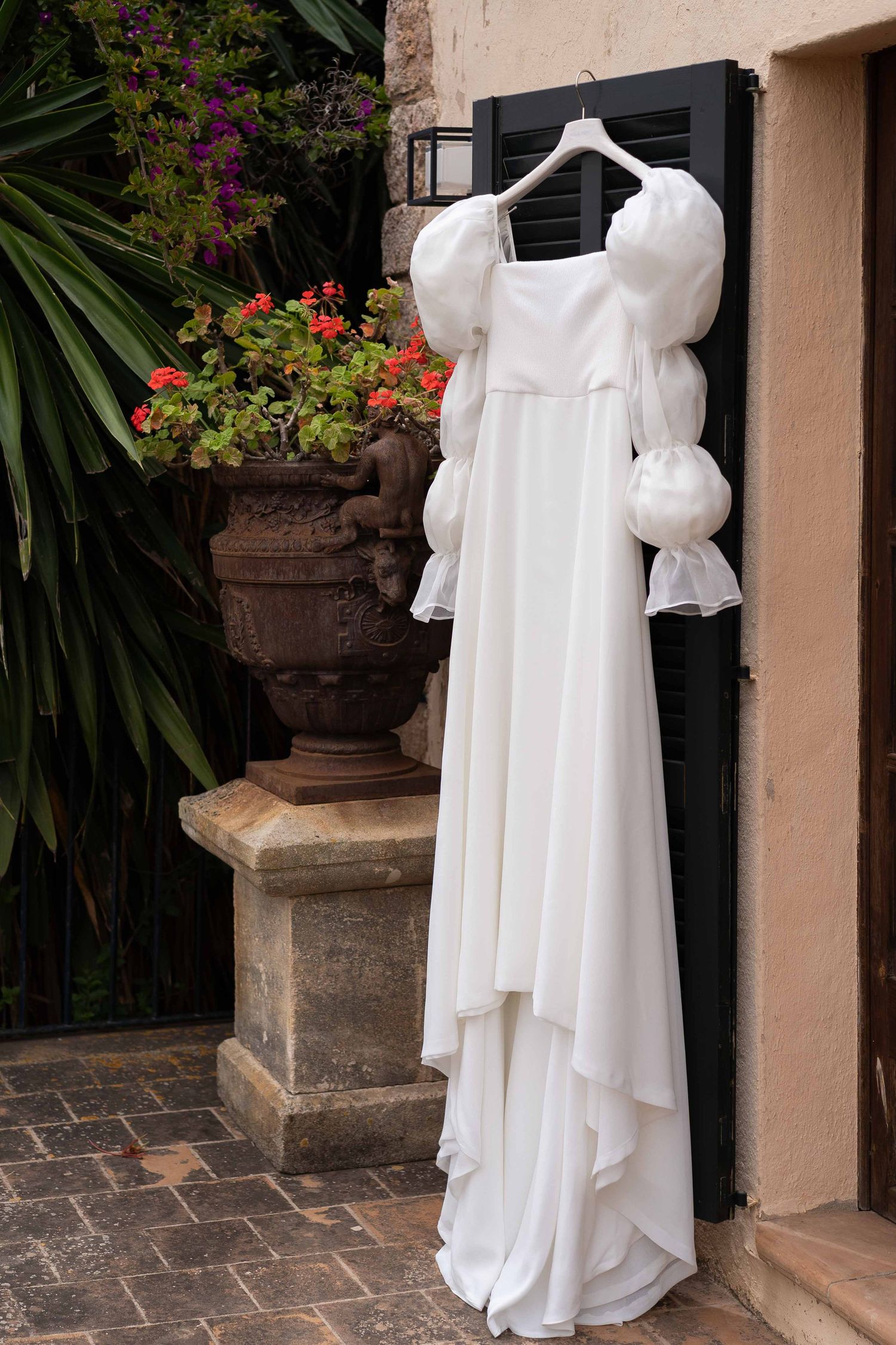 White wedding dress displayed on hangers against a Mediterranean villa wall with potted flowers.
