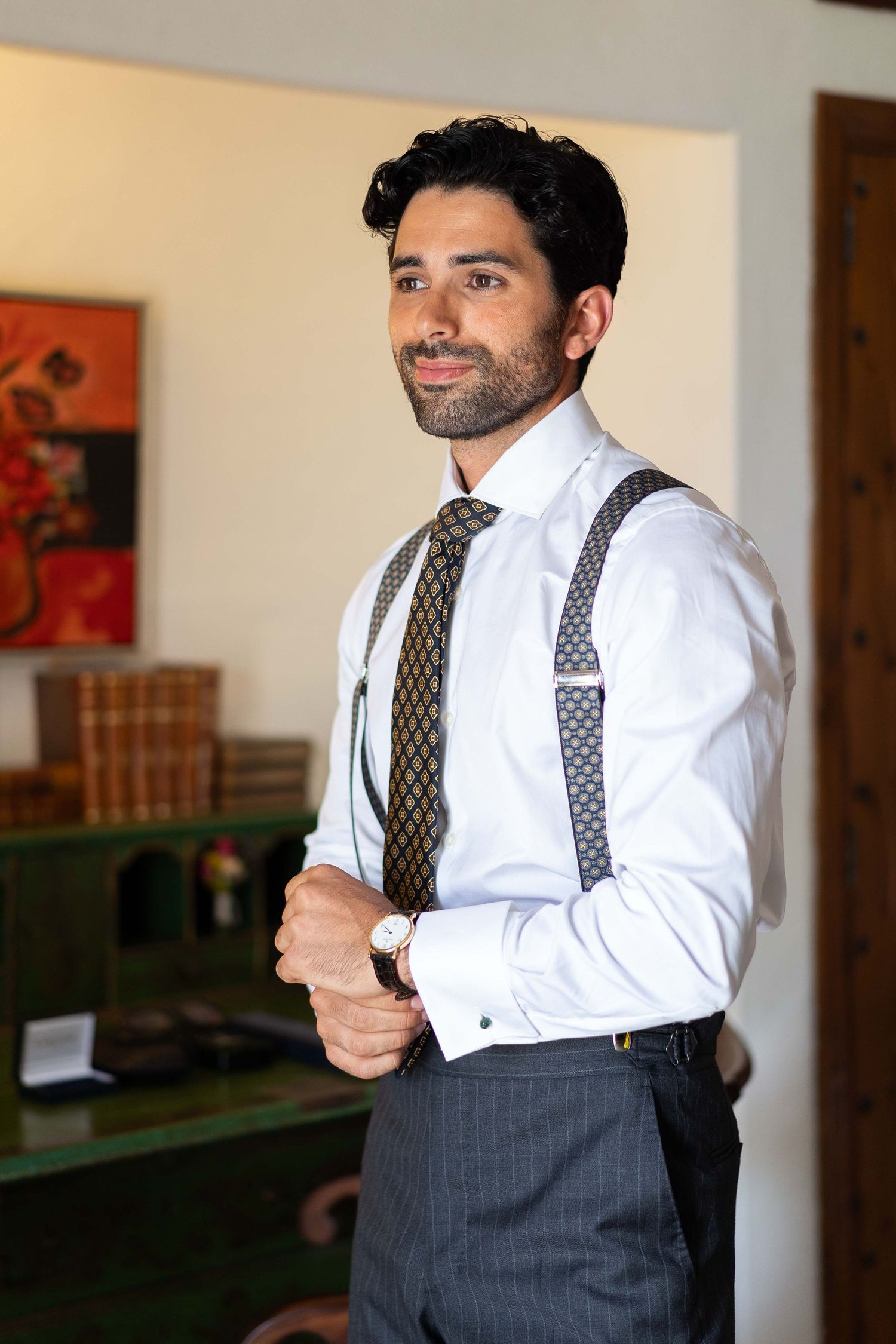 Man in formal attire with suspenders and patterned tie adjusting his outfit indoors.