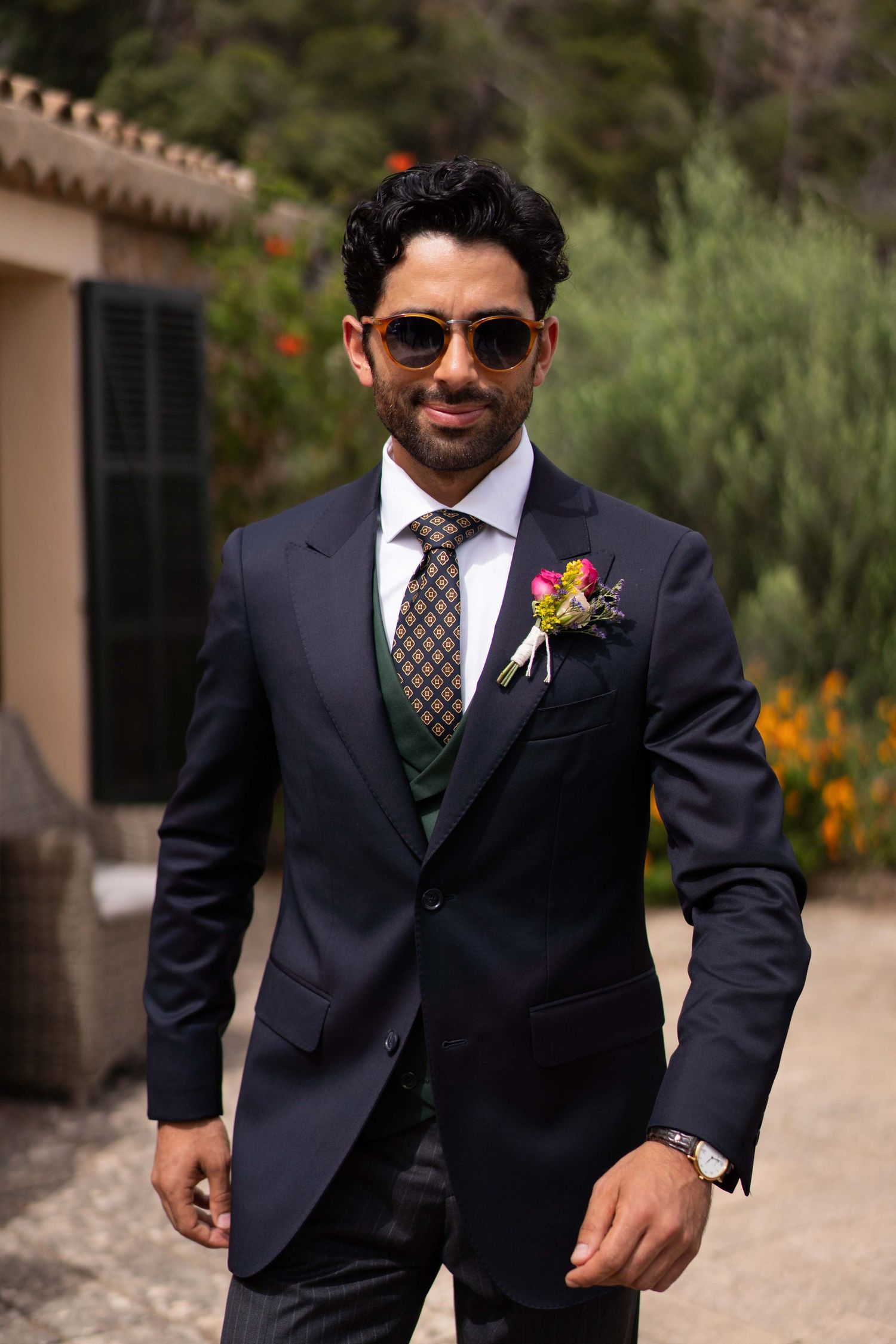 Person in formal dark suit with boutonniere and sunglasses at outdoor wedding venue.
