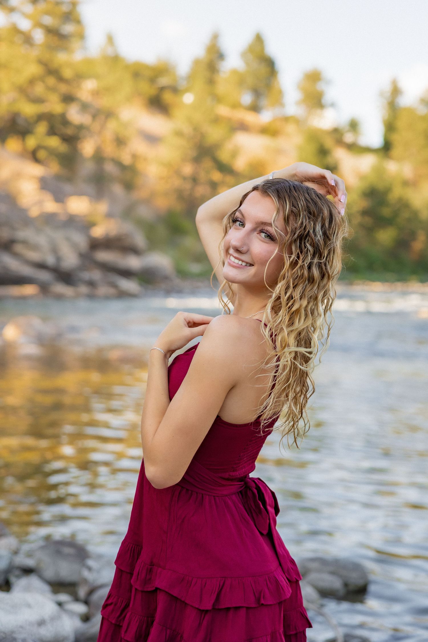 A person in a burgundy dress poses by a river at sunset with rocks and trees in the background.