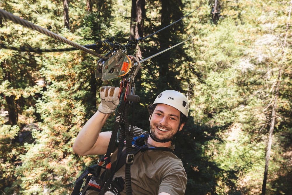 Adventurers smiling and having fun while ziplining between tall trees in a sunlit forest.