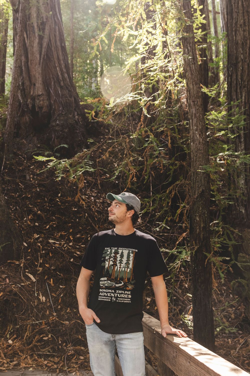 Person in a baseball cap and t-shirt exploring a scenic redwood forest trail on a sunny day.