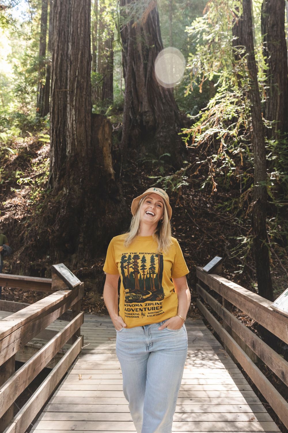 A person in a yellow shirt and hat poses on a wooden boardwalk surrounded by towering redwood trees.