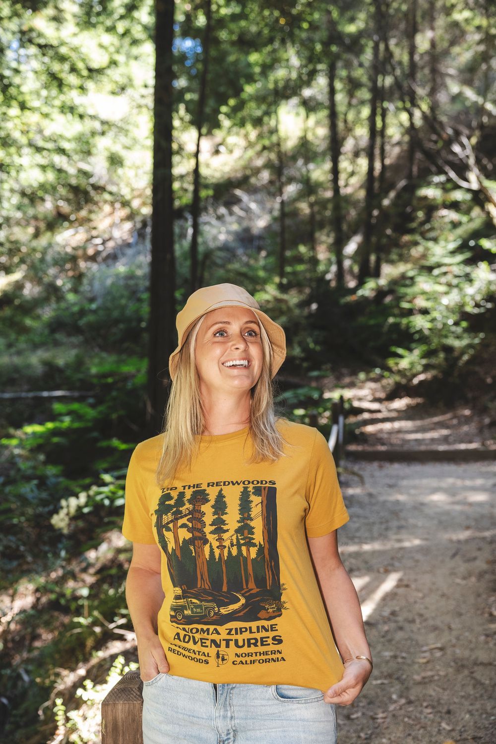 A hiker in a yellow graphic t-shirt and sun hat smiles while exploring a sunlit forest trail.