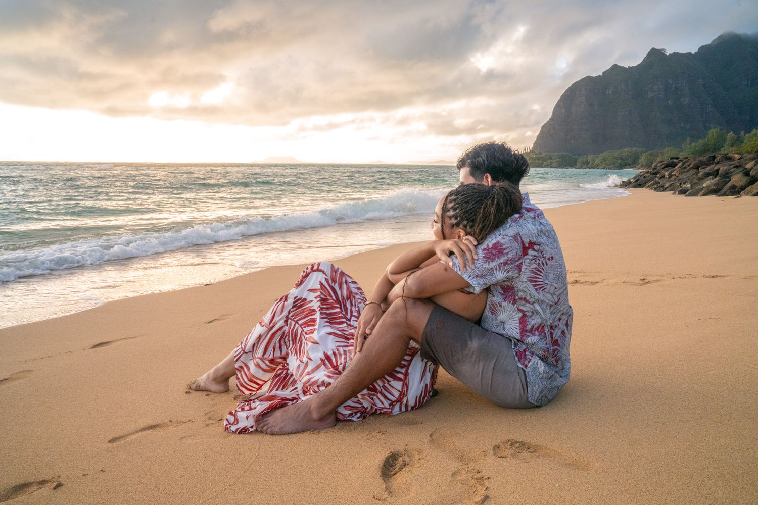 A couple relaxes together on a sandy beach at sunset with mountains in the background.