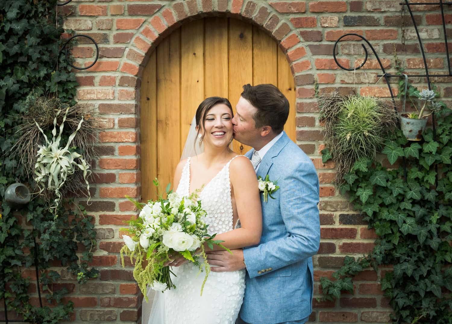 A romantic wedding photoshoot against a rustic brick wall with climbing ivy vines and warm lighting.