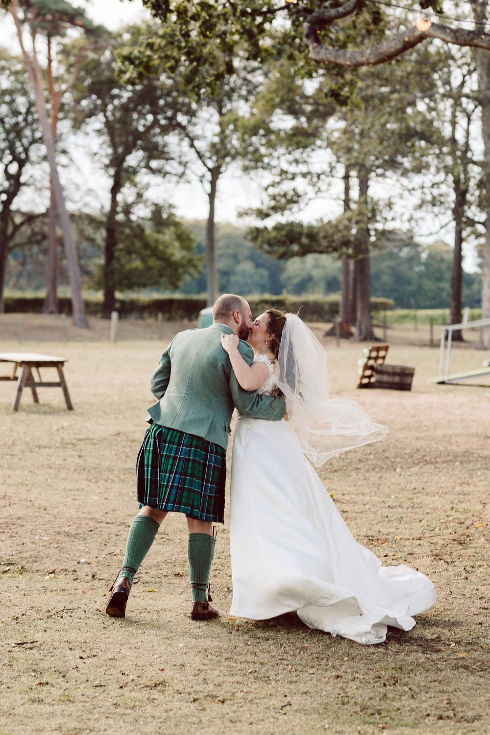 A couple in wedding attire dances and spins across a grassy outdoor venue with pine trees in the background.