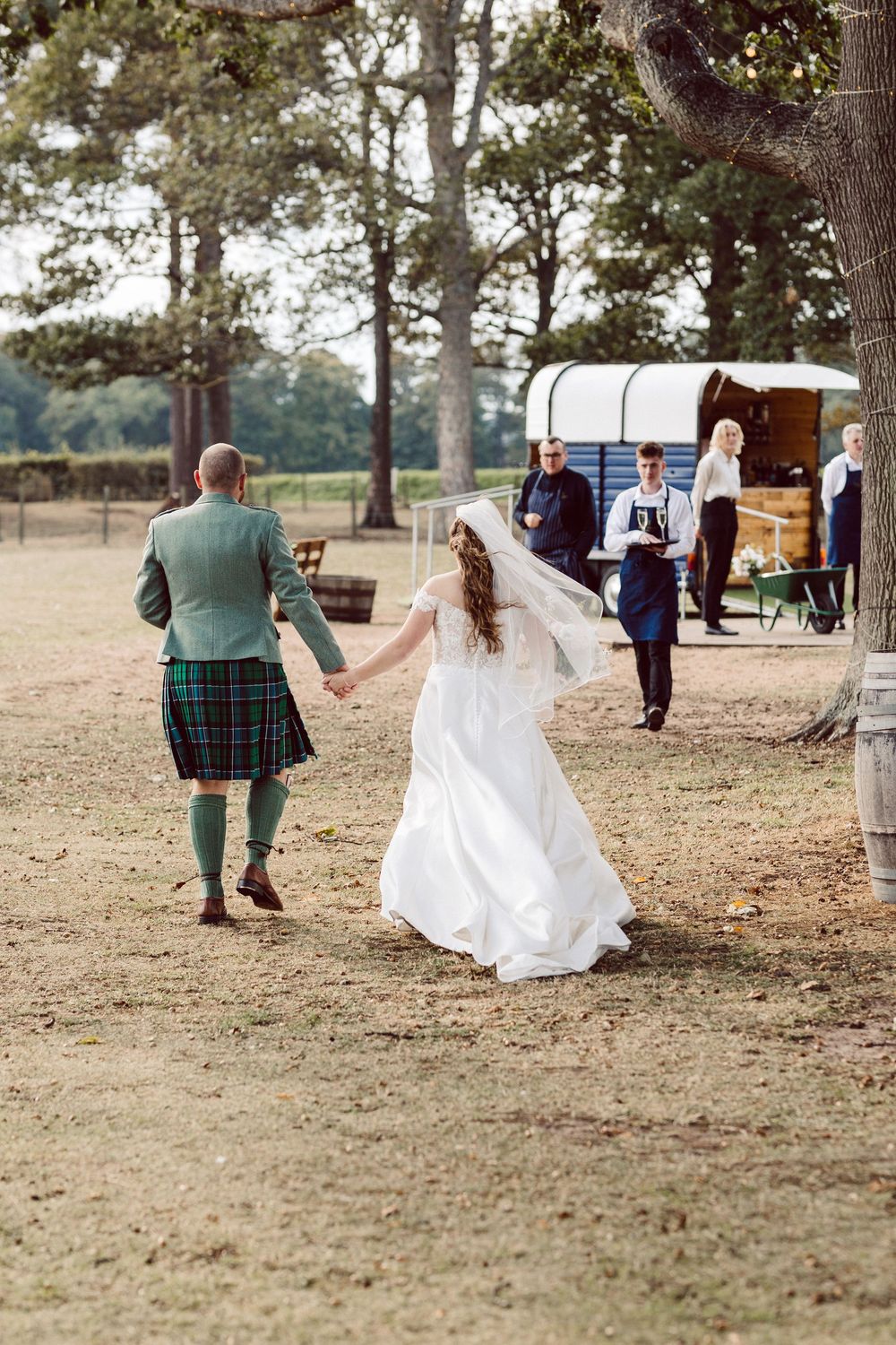 A wedding party walks across a grassy field with trees in the background, showing movement and celebration.