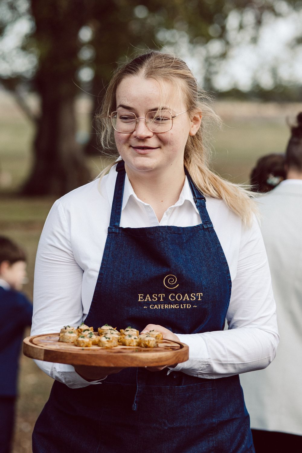 Server in navy apron and white shirt carries wooden serving boards with appetizers outdoors at catering event.