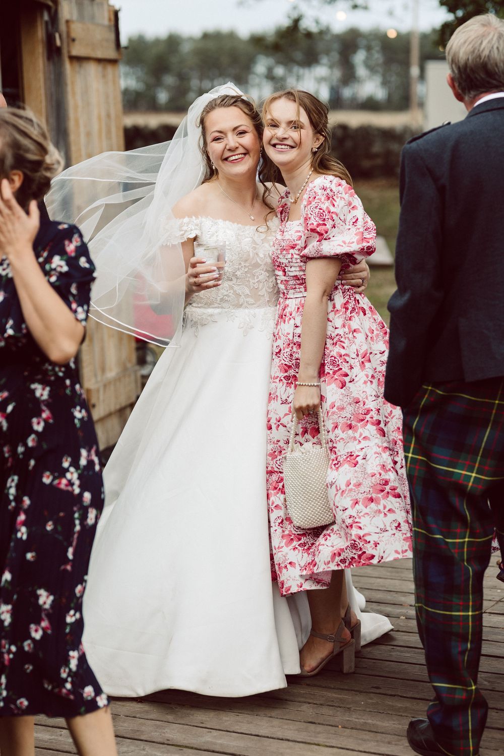 A bride in a white lace gown stands with a guest in a pink floral dress at an outdoor wedding reception.