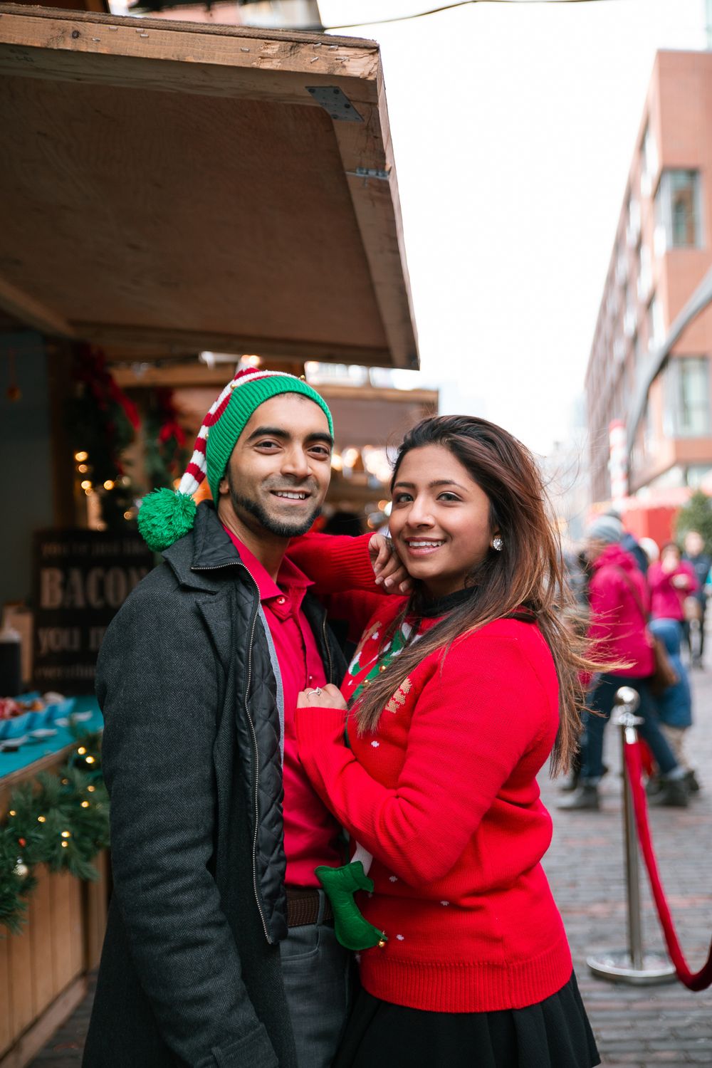 Smiling couple in festive holiday sweaters posing at a Christmas market stall.