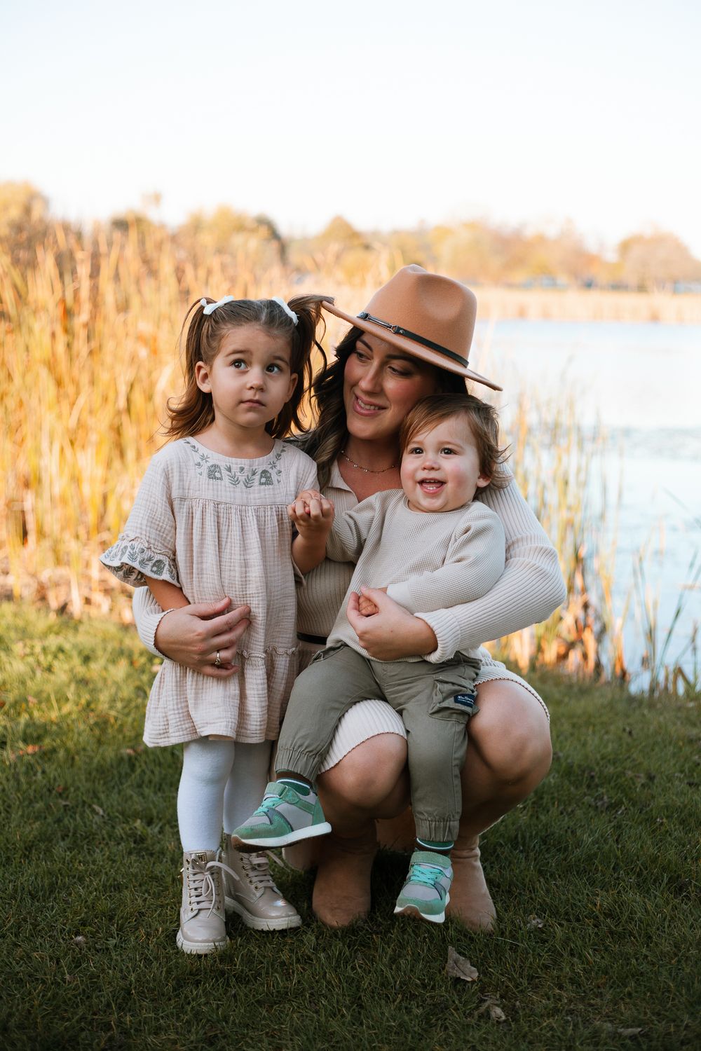 Smiling mother in tan hat crouching with two young children near autumn lakeside.