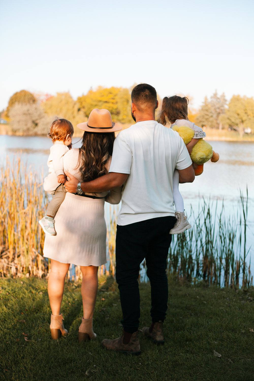 Family of four viewed from behind near a lake, parents holding two young children in fall setting.