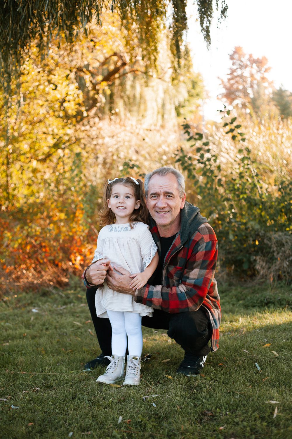 Smiling man in red plaid jacket crouching with young girl in white dress in autumn park.