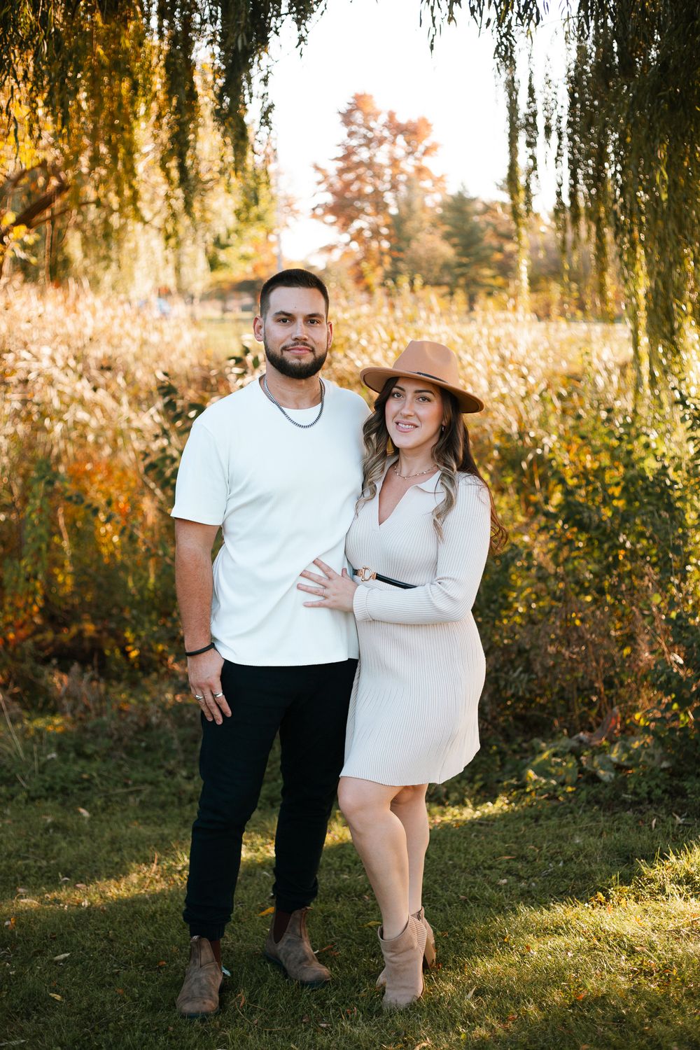 Couple posing outdoors in autumn, man in white tee and black pants, woman in beige dress and tan hat.