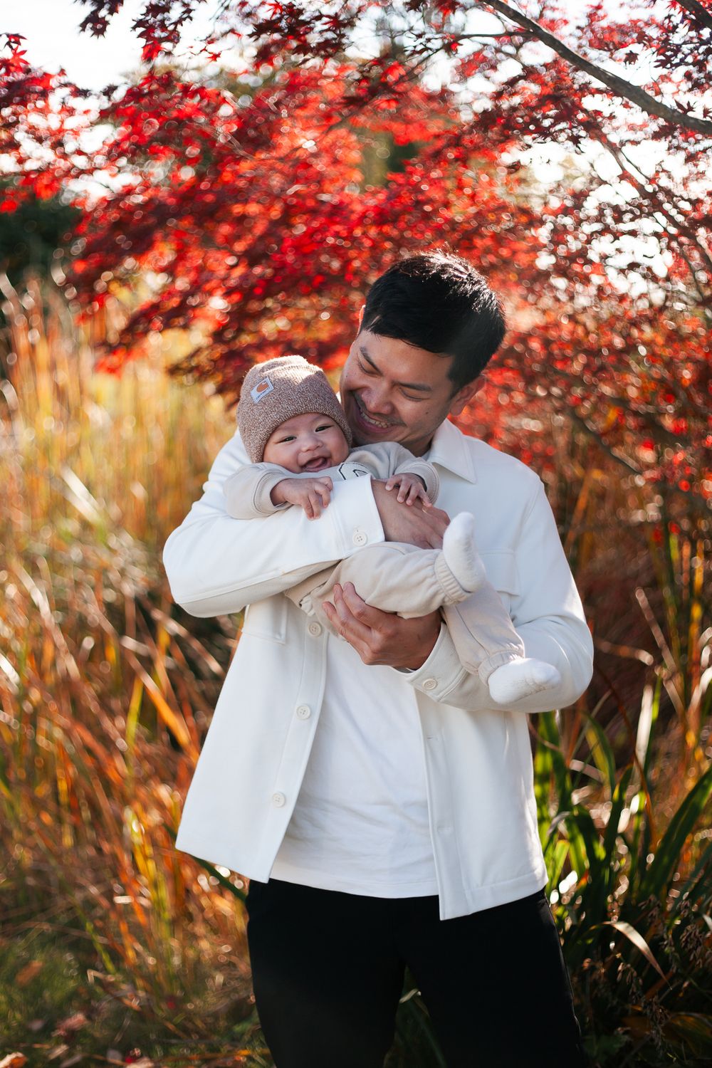 Happy father in white shirt holding laughing baby outdoors among vibrant red foliage.