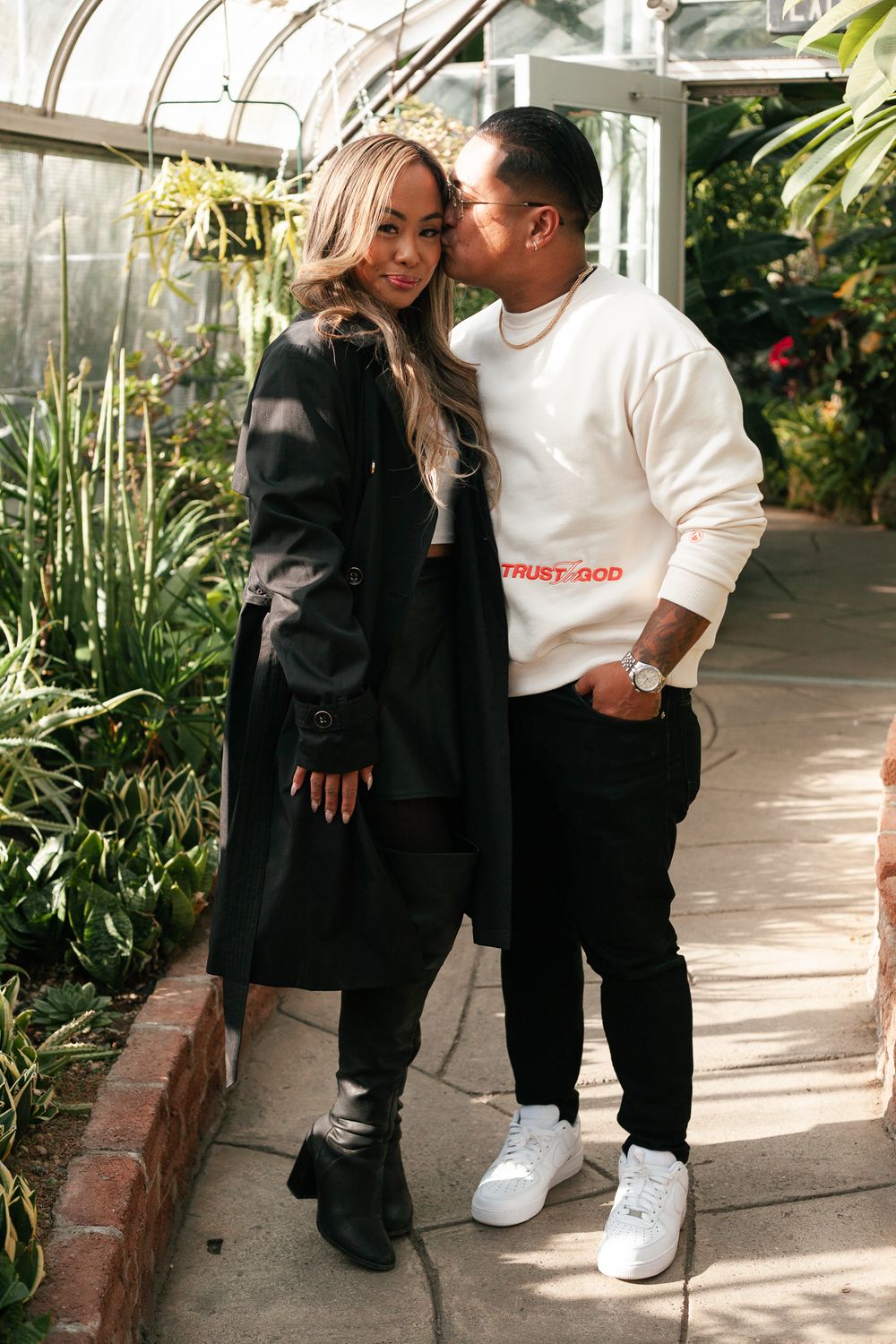 Couple posing in greenhouse, man kissing womans head, both wearing stylish black outfits.