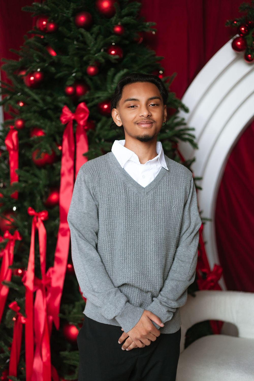 Young man in gray V-neck sweater smiling before red-decorated Christmas tree.