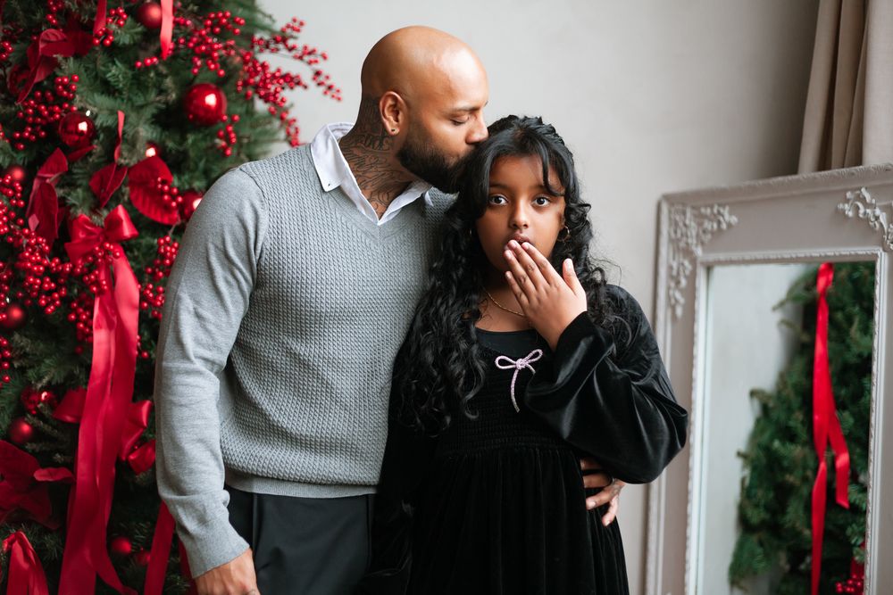 Bald man kissing young girl on head near decorated Christmas tree indoors.