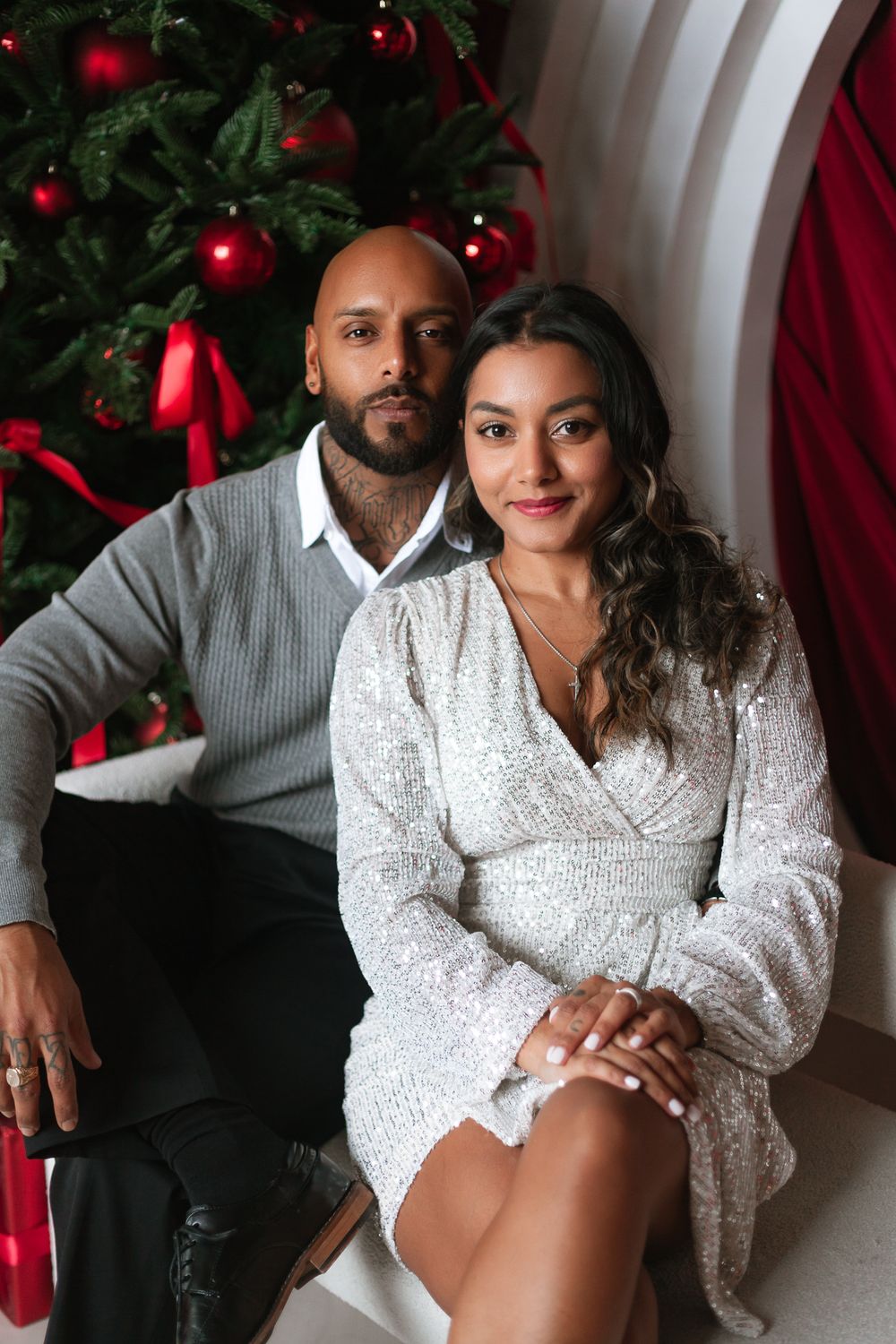 Couple posing by Christmas tree, man in gray sweater, woman in silver sequin dress.