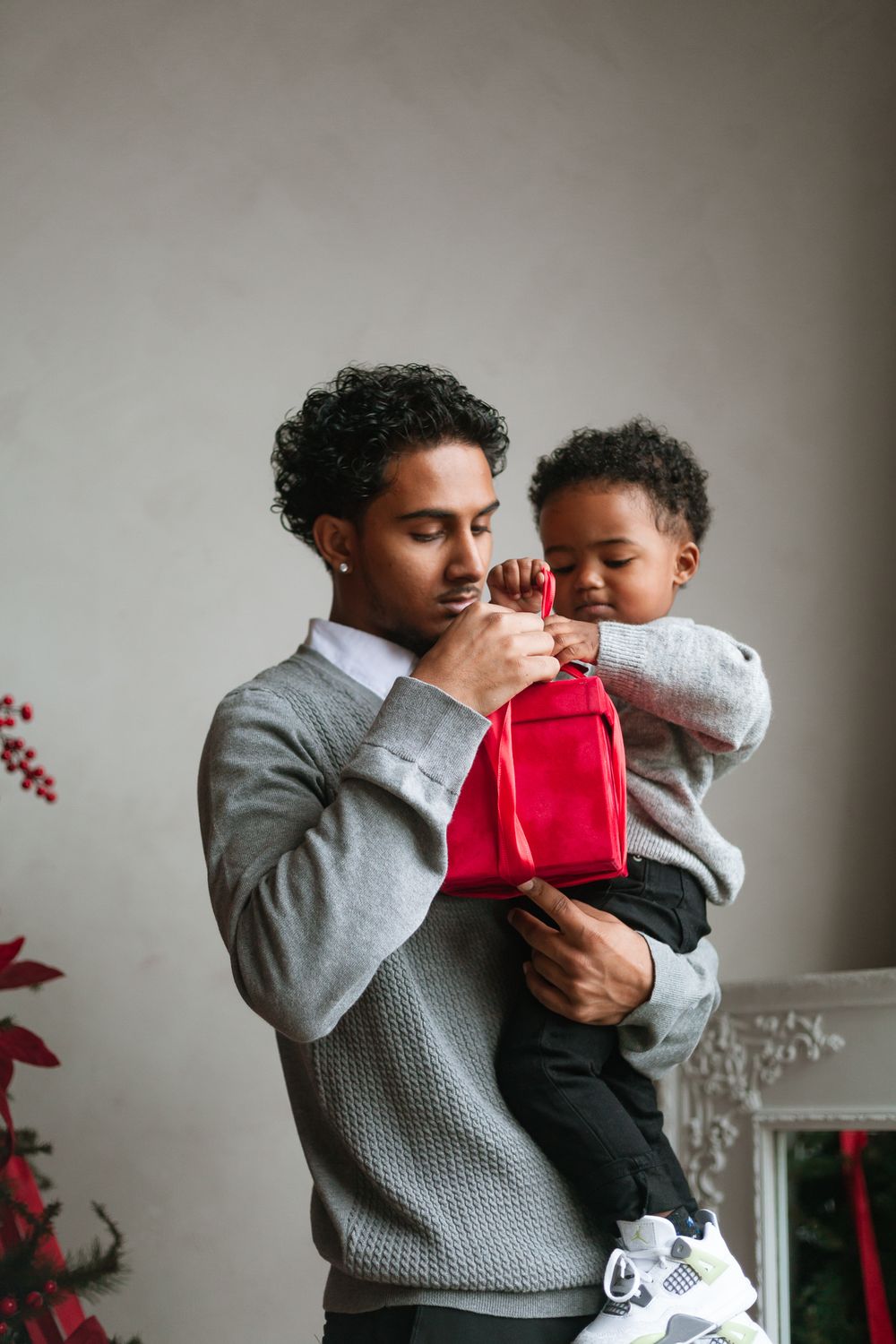 Young man in gray sweater holding toddler with red gift bag near holiday decor.