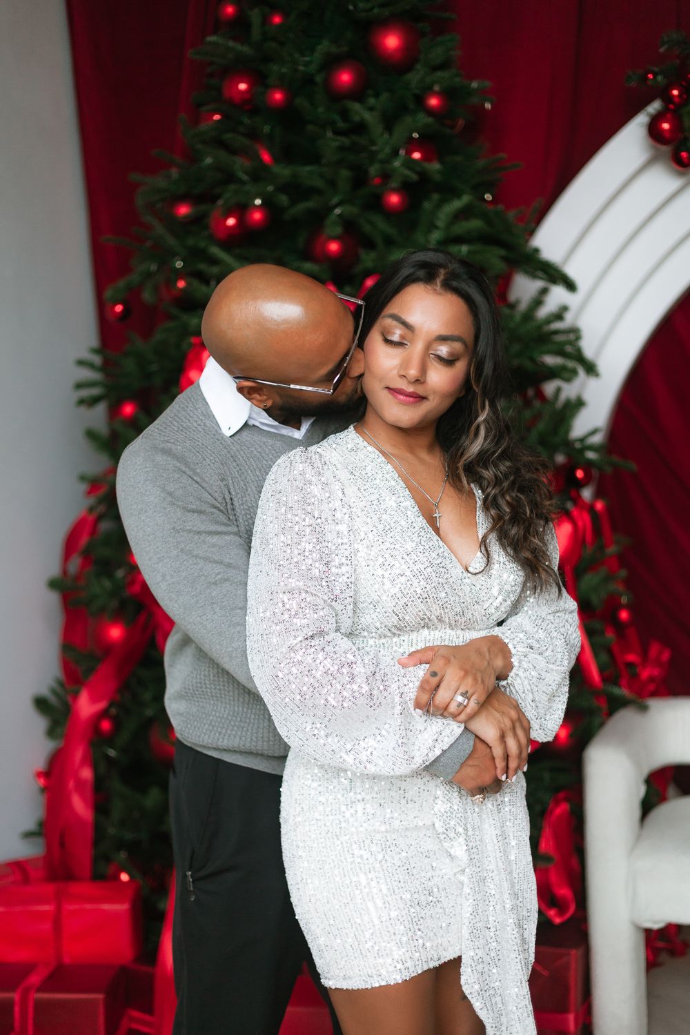 Couple embracing before Christmas tree, man in gray sweater hugging woman in silver sequin dress.