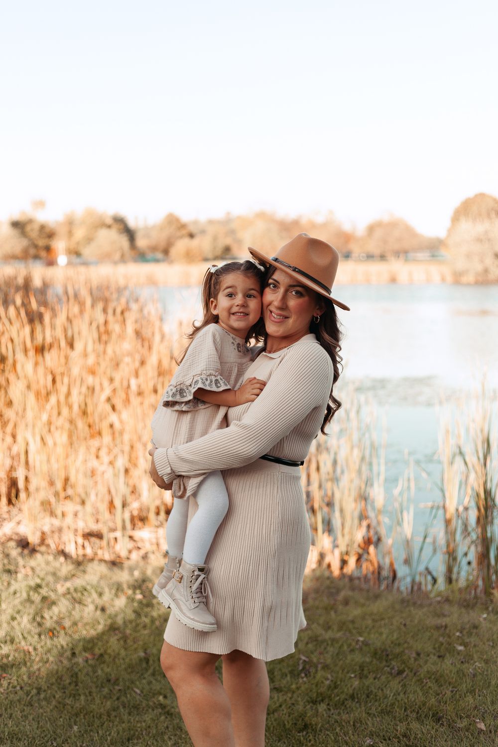 Smiling mother in tan hat and beige dress holding young daughter near autumn lakeside.
