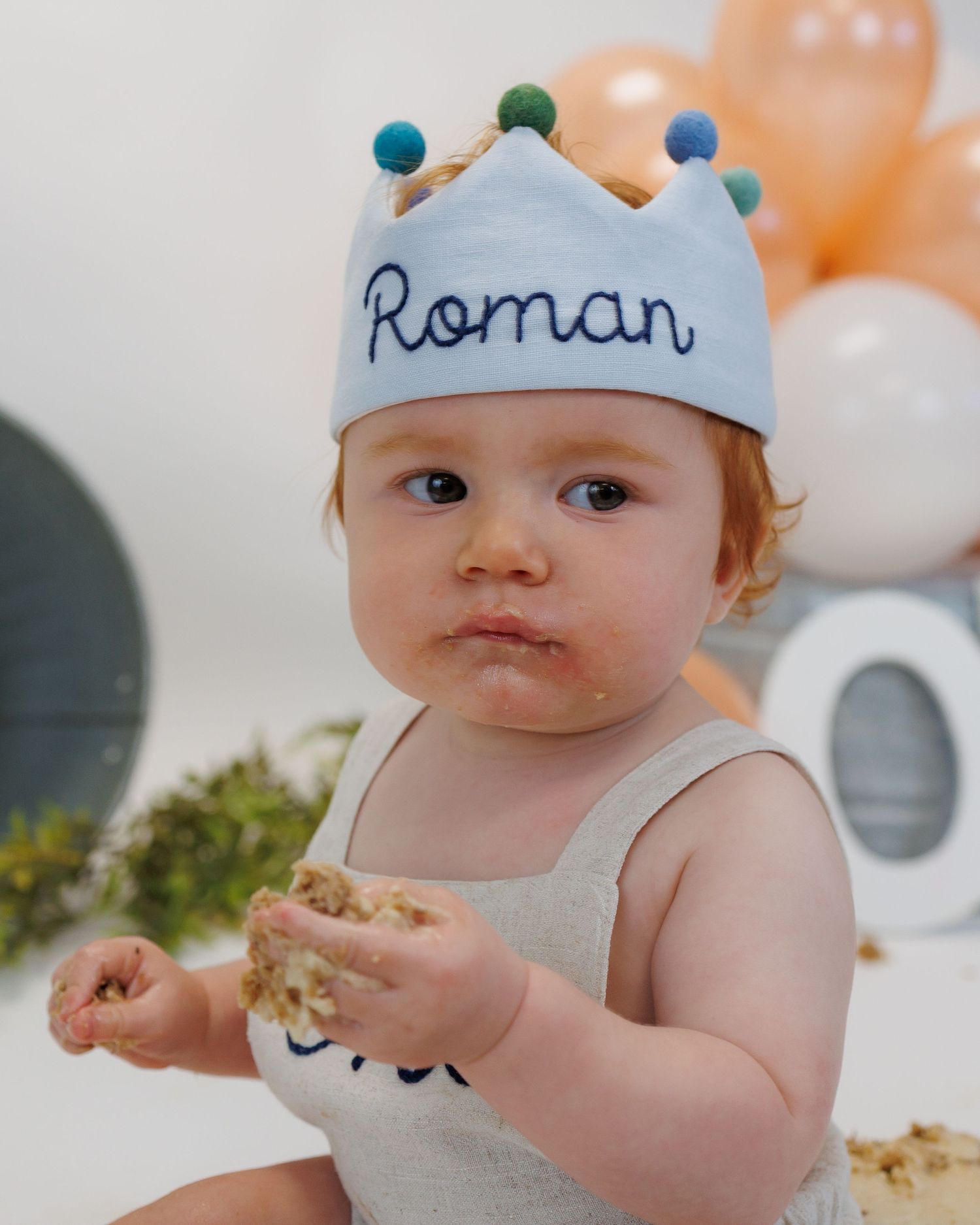 Baby wearing blue paper crown sits with cake smash setup in peach and white themed first birthday celebration.