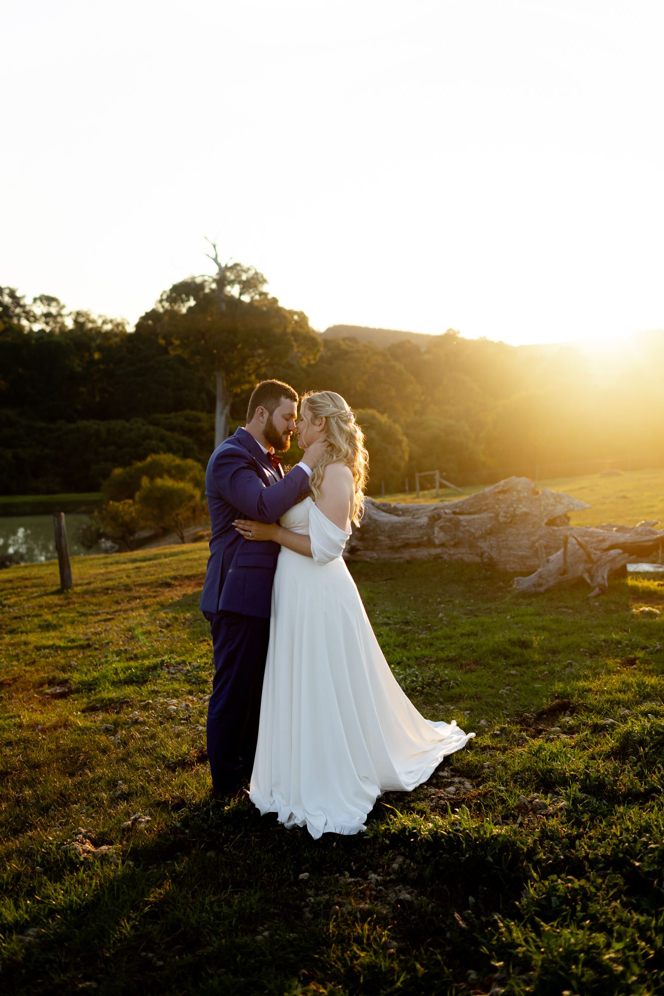 A couple embraces outdoors in wedding attire, with the bride in white lace and groom in navy suit on a sunny garden day.