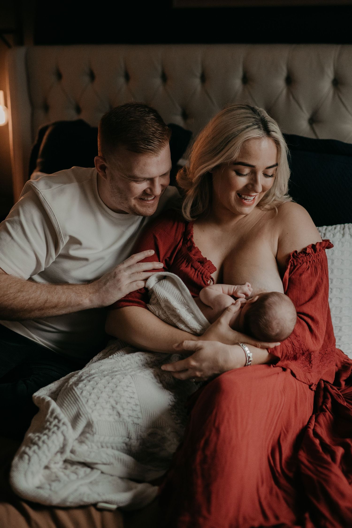 A couple lying on a bed, smiling at their new born baby while the baby nurses in his mother's arms.