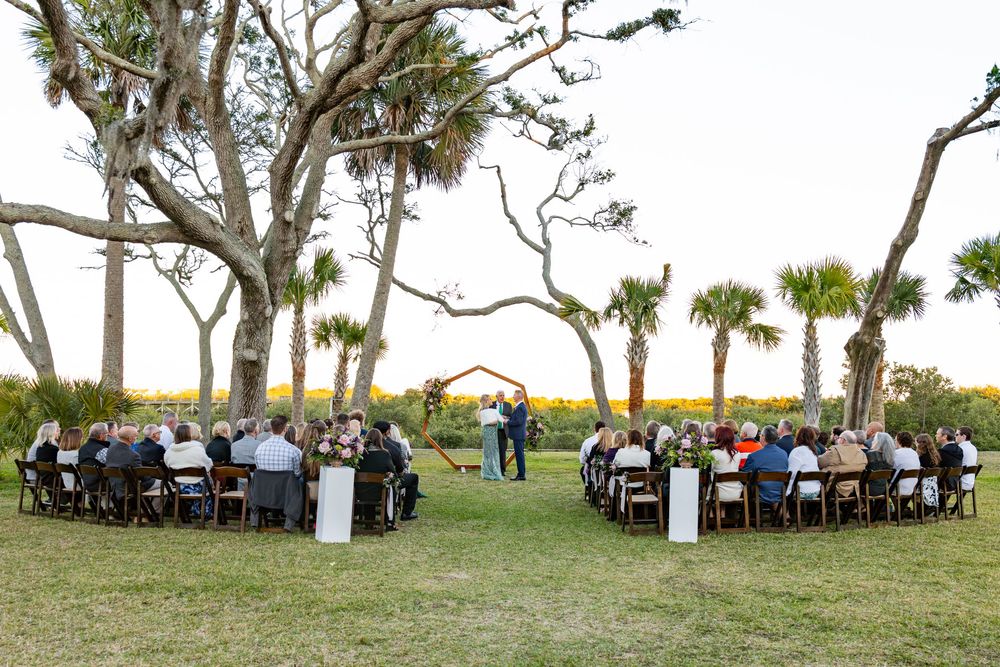 Outdoor wedding ceremony setup with white chairs arranged under oak trees beside a coastal marsh at sunset.