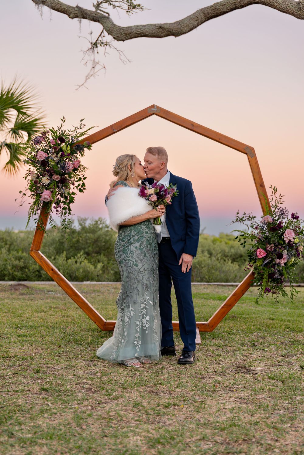 A couple shares a romantic moment at sunset under a hexagonal wooden arch decorated with pink and purple florals.