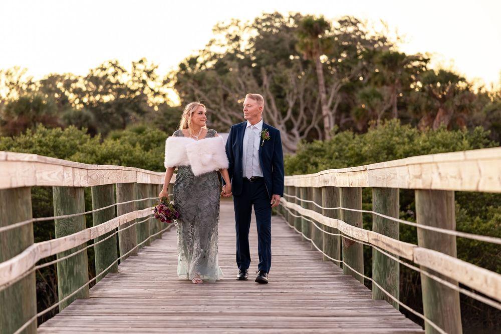 Couple walking together on a wooden boardwalk surrounded by coastal vegetation at sunset.