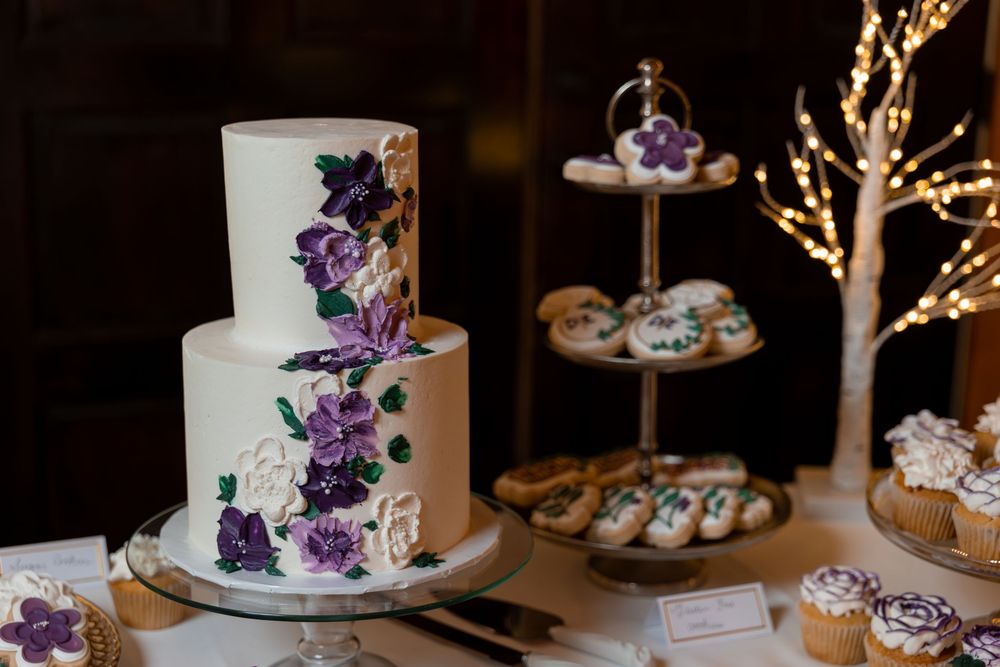 White wedding cake adorned with cascading purple orchids and delicate gold branches on an elegant dessert table display.