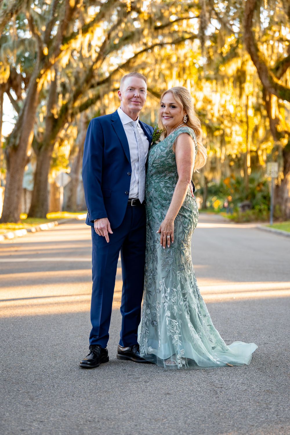 A couple poses together on a tree-lined path during golden hour sunset with Spanish moss hanging overhead.