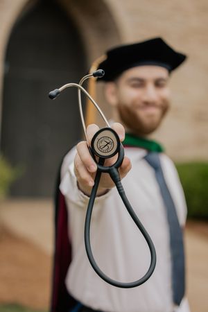 Smiling male medical graduate in cap and gown holding a stethoscope toward camera outdoors.
