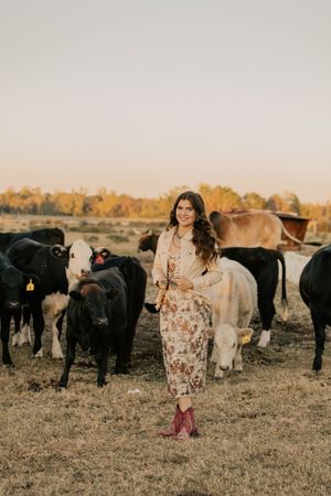 Smiling woman in floral dress and red boots standing among cattle on a farm at golden hour.