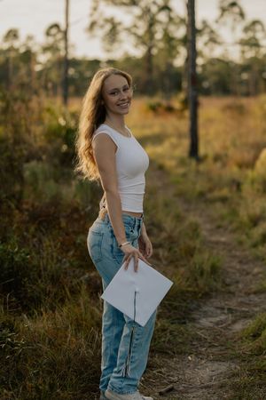 Young woman in white crop top and jeans holds graduation cap while smiling outdoors in golden hour light.