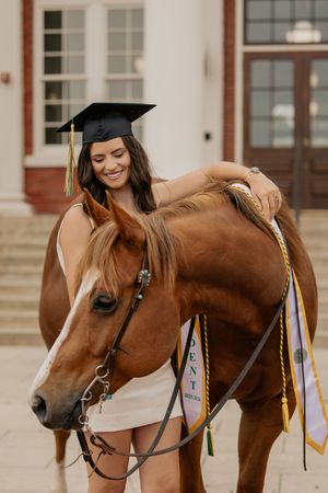 Smiling female graduate in cap and gown poses with a chestnut horse outside a brick building.